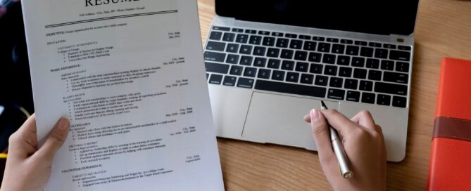A person holding a paper resume while seated at a laptop computer.